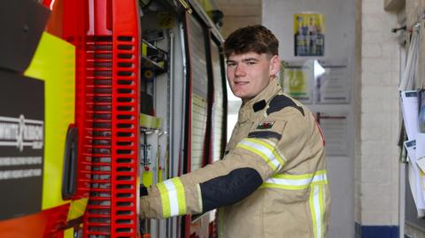 Bobi in a firefighter uniform looking at the camera and holding on to the side of a fire engine. He has short dark hair and is clean shaven. 